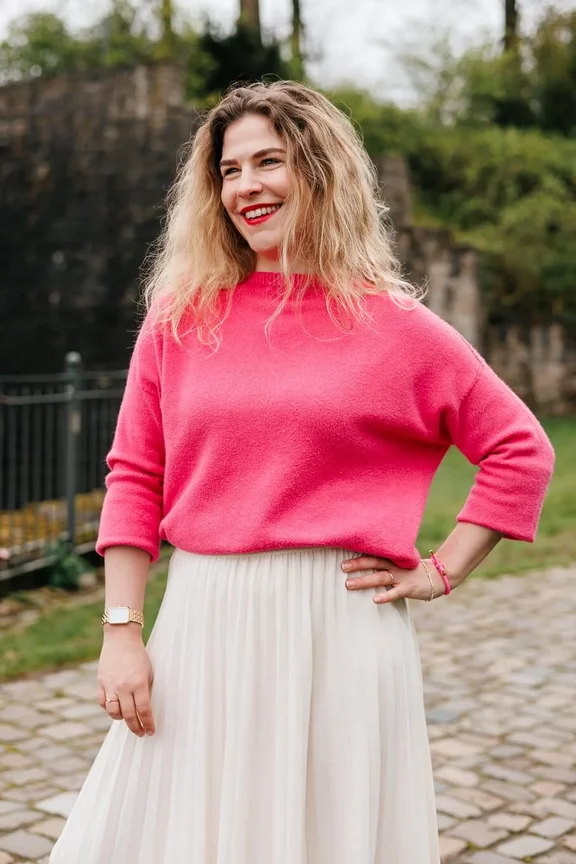 Eine Frau mit lockigem blondem Haar trägt einen rosa Pullover und einen weißen Rock und steht draußen vor einer Steinmauer mit Grün im Hintergrund.
